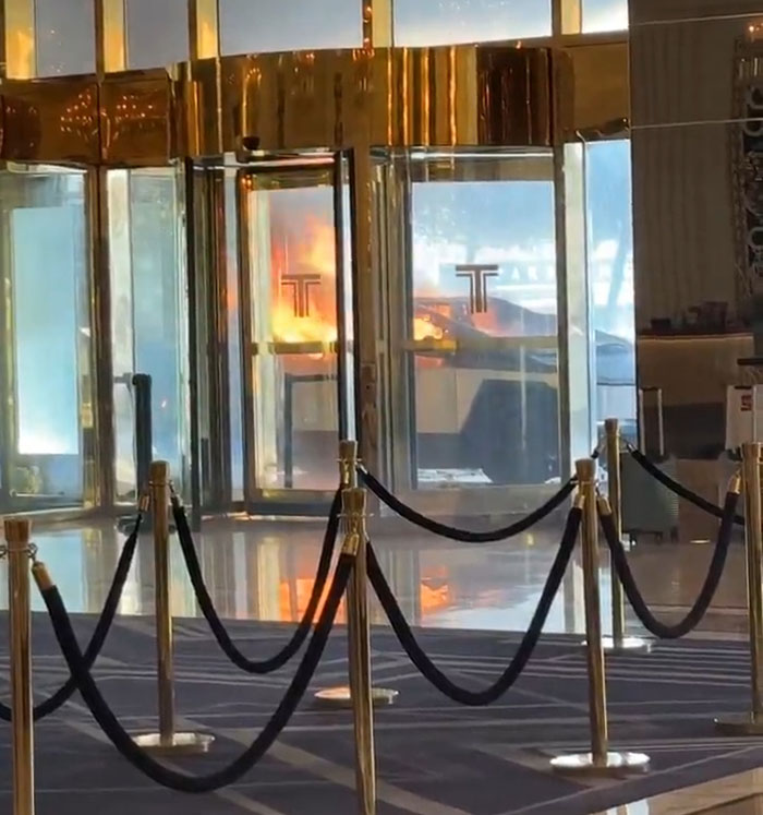 Cybertruck surrounded by flames in a building entrance, reflecting off golden glass doors.