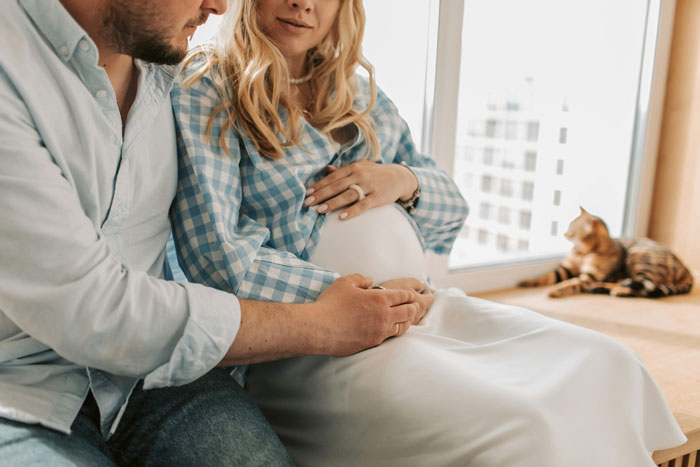 Dad and expectant mother sitting together, hands on her belly, with a cat by the window.