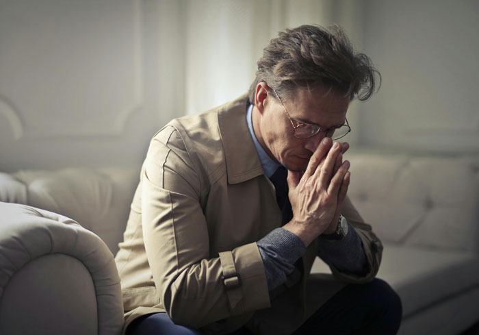 Man on a sofa showing dad grief, wearing a beige coat and glasses, with his hands clasped in contemplation.