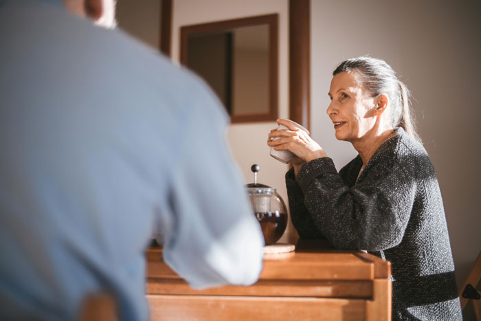 A woman in a sweater, seated at a table with a coffee pot, talking to a man, capturing a moment of connection.