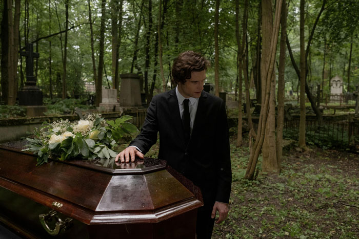 A man in a suit mournfully stands by a casket in a wooded cemetery, symbolizing dad grief.