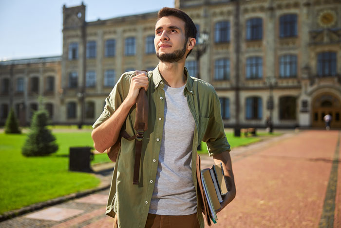 Male student outside a university building, carrying books, related to college funding challenges.