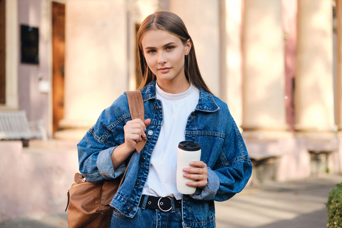 Young woman in denim jacket, holding a cup, symbolizes struggles with college costs compared to lavish sibling lifestyle.