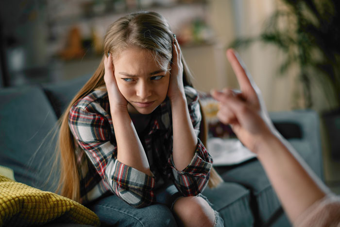 Teen girl covering ears, looking frustrated while sitting on a couch.