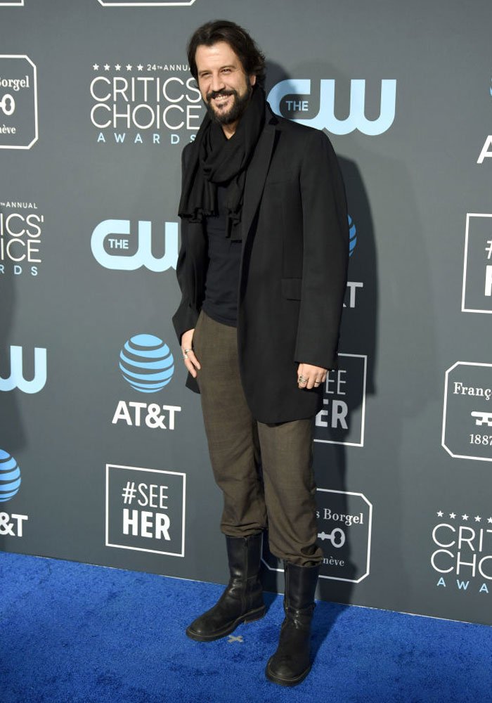 Stefan Kapičić at Critics' Choice Awards, related to Deadpool cast, stands on blue carpet in black coat and scarf.