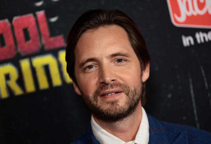 Actor Aaron Stanford at Deadpool event, wearing a blue suit, posing in front of a movie-themed backdrop.