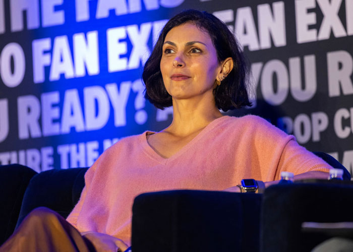 Actress Morena Baccarin in a pink sweater sitting at a Deadpool cast panel, with promotional text background.