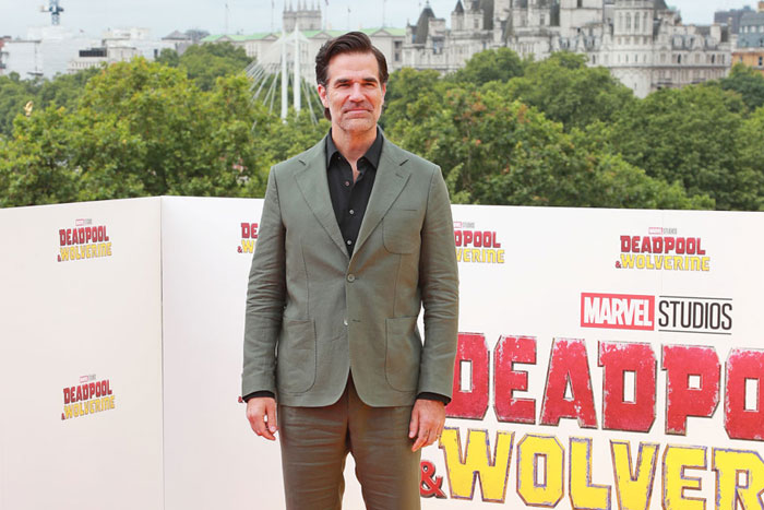 Actor Rob Delaney poses at a "Deadpool & Wolverine" event, wearing a green suit, standing in front of branded backdrop with trees behind.