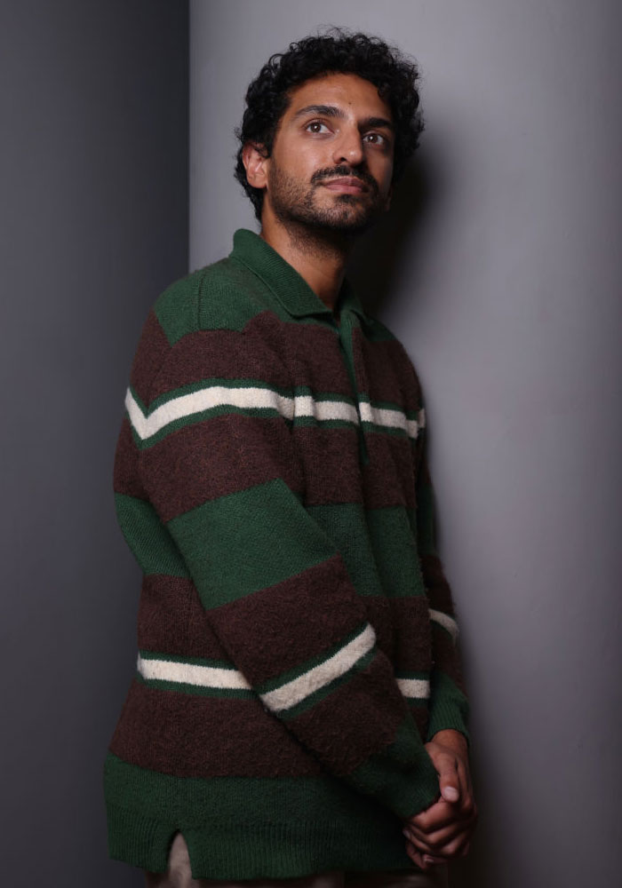 Actor Karan Soni in a green and brown sweater, likely part of the Deadpool cast, standing against a gray wall.