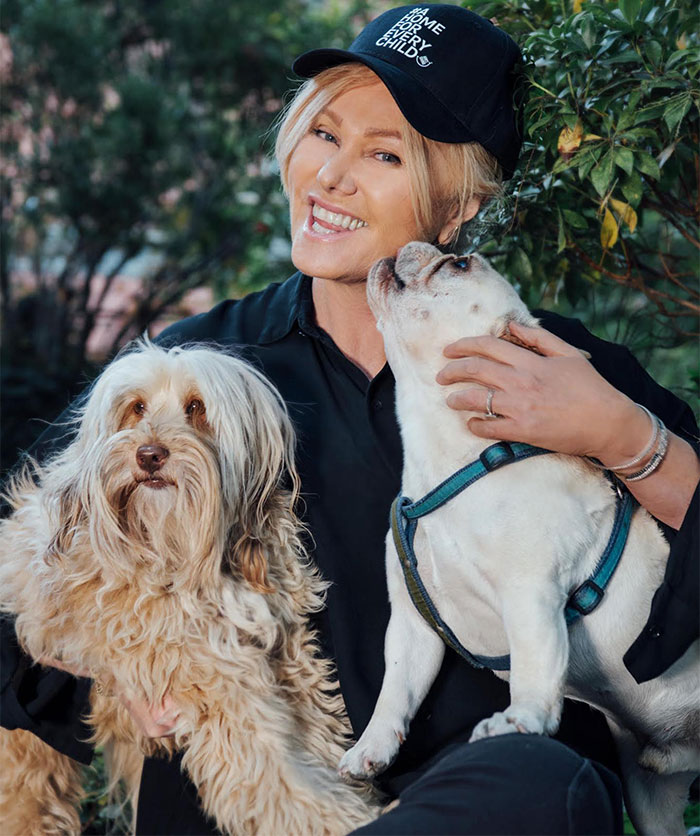 Woman smiling with two dogs in a garden, wearing a hat.