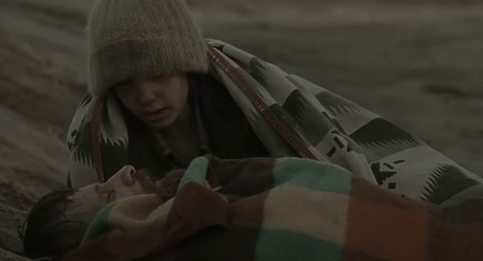 Child comforting an adult wrapped in blankets on a deserted landscape, capturing a sad and depressing movie ending.