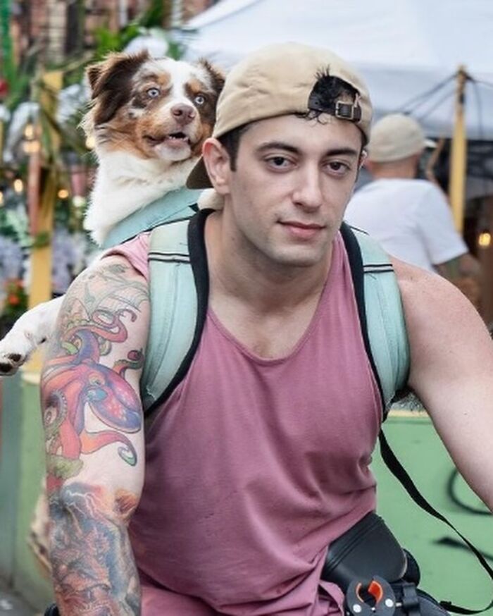 Tattooed man with NYC dog in backpack, both gazing ahead in a lively street scene.