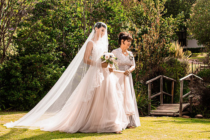 Bride walking with her mom in a garden, both in elegant dresses, highlighted by natural sunlight.