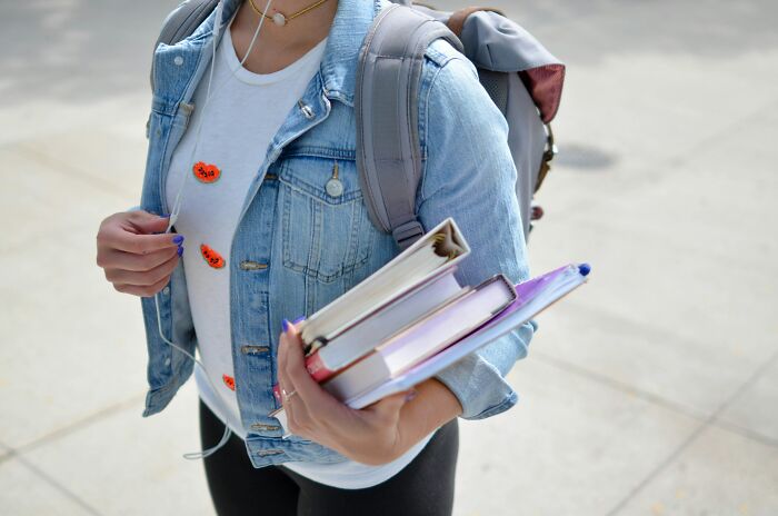 Person in denim jacket holding books and wearing a backpack, possibly discussing the biggest scam in human history.