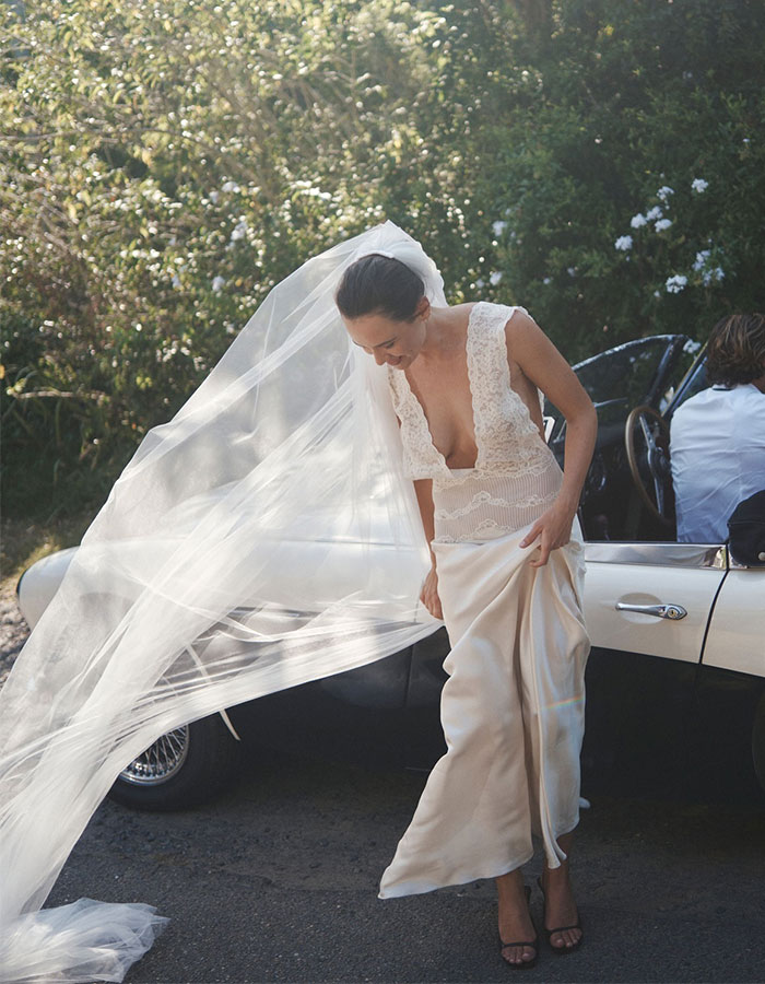 Australian bride in a wedding dress with a lace top and flowing skirt, standing by a white car with her veil blowing in the wind.