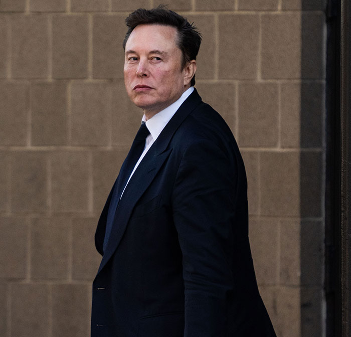 Man in a suit standing against a brick wall background, related to discussion about influence and wealth.