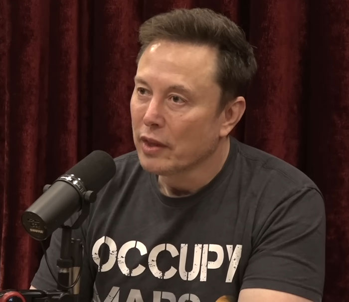 Man in "Occupy Mars" shirt, speaking into a microphone, against a red curtain backdrop.