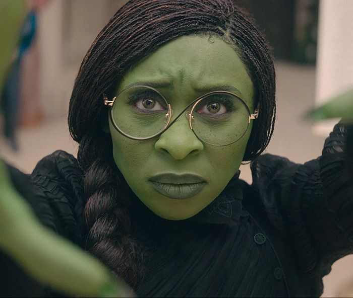 Green-skinned character in glasses and braided hair from 'Emilia Pérez'.