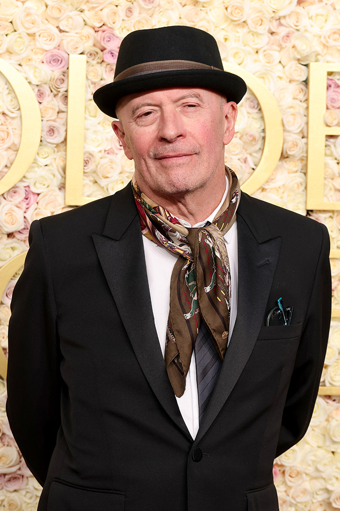 Director in a formal suit and hat at an event, against a floral backdrop.