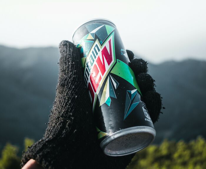 Gloved hand holding a Mountain Dew can with a scenic mountain background.