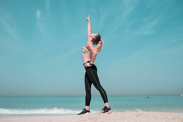 Person in athletic wear stands on beach, pointing upward under a clear sky.