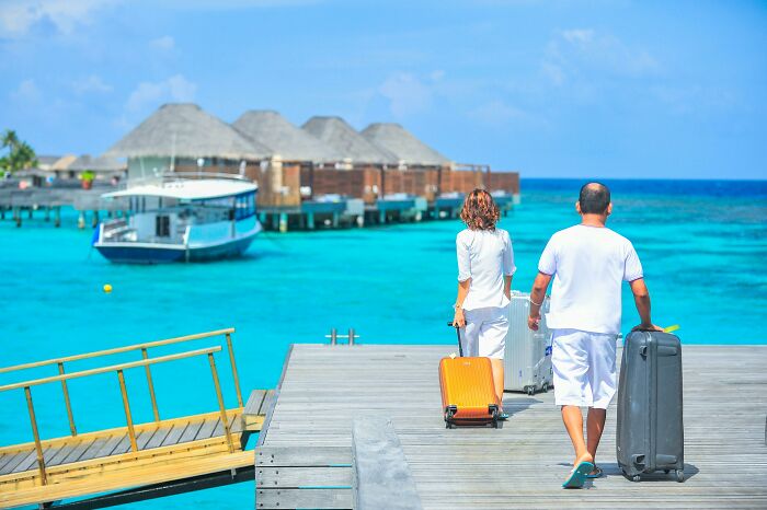 Couple with suitcases on a tropical pier, reflecting luxury travel choices affected by cost.