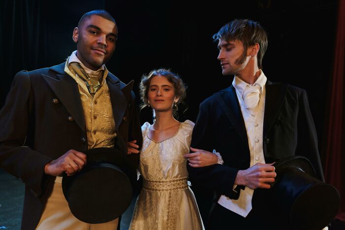 Three people in period costumes holding top hats, portraying a scene.