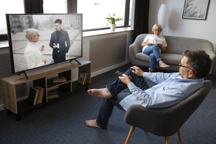 A man watching TV with a remote, while a woman reads on a couch.