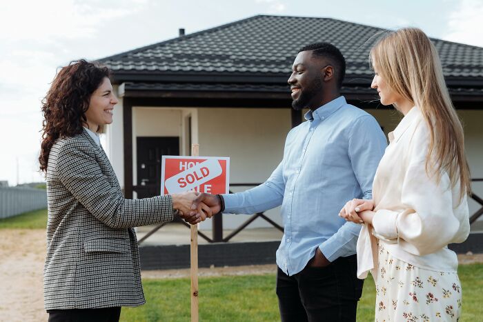 Real estate agent closing deal with couple, standing by "Sold" sign in front of house, symbolizes cost-conscious buying.