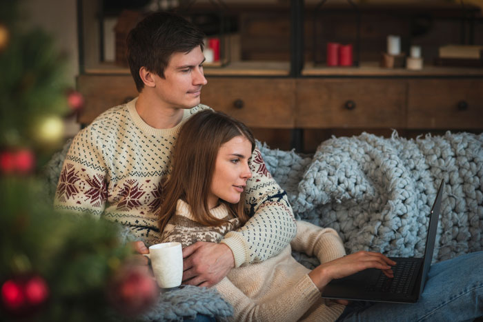 A couple on a sofa, wearing winter sweaters, with a laptop and mug, depicting family New Year's Eve drama.