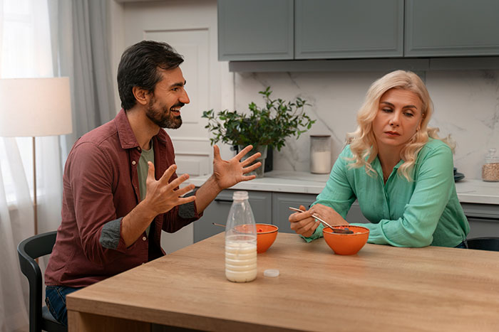 Husband speaking animatedly, wife looking away, sitting at a kitchen table with orange bowls and a milk bottle.