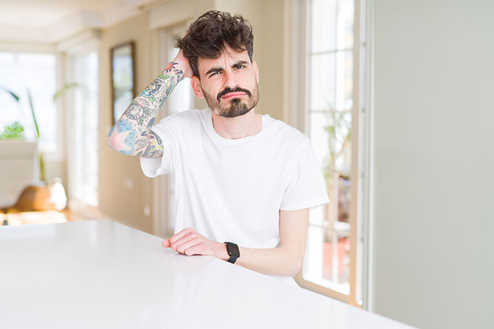 Man in a white shirt with tattoos looking puzzled, in a bright room, related to in-laws' anniversary collage discussion.