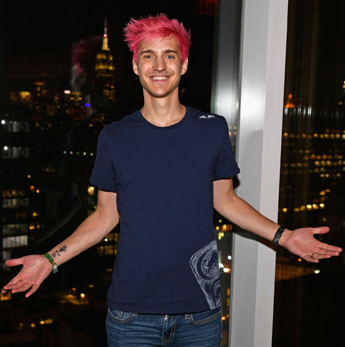 Streamer Ninja (Richard Tyler Blevins) with pink hair, wearing a navy shirt, stands with arms open against a cityscape backdrop.