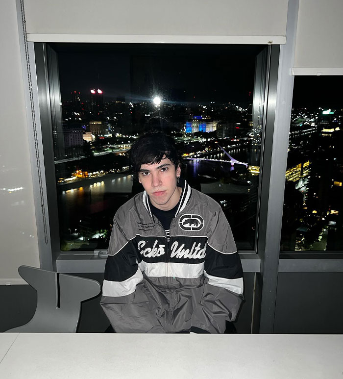 Spreen (Iván Buhajeruk) sits in front of a city nightscape, wearing a dark and white jacket in a high-rise room.