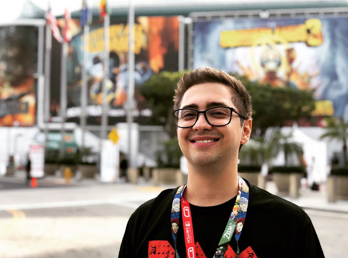 Smiling Alanzoka (Alan Ferreira Pereira) with glasses at an outdoor event, wearing a lanyard, representing famous streamers on Twitch.