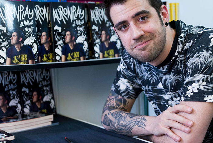 Twitch streamer Auronplay (Raúl Álvarez Genes) sitting at a table with books in the background, wearing a floral shirt and showing arm tattoos.