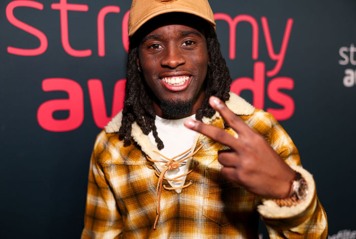 Famous streamer Kai Cenat in a plaid jacket and cap, smiling and gesturing peace sign at an awards event.