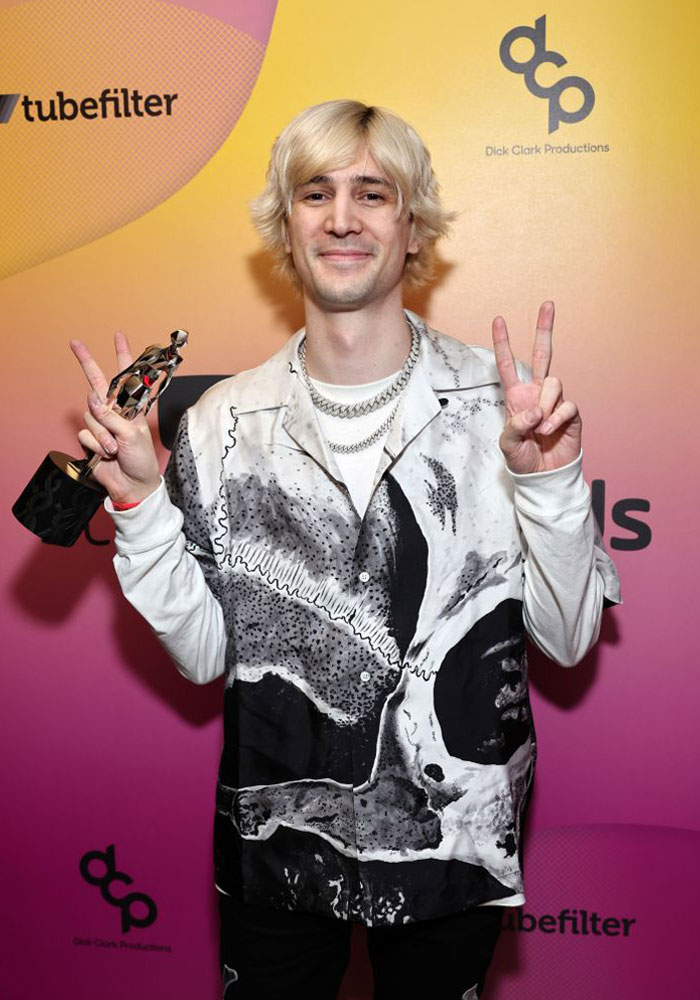 Streamer xQc (Félix Lengyel) holding an award, wearing a patterned shirt, poses with peace signs.