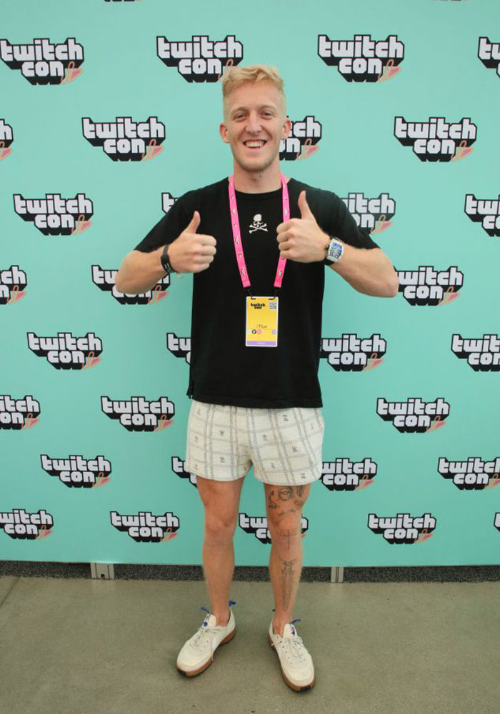 Famous streamer Tfue (Turner Tenney) at TwitchCon, smiling and giving thumbs up, standing against a Twitch-themed backdrop.