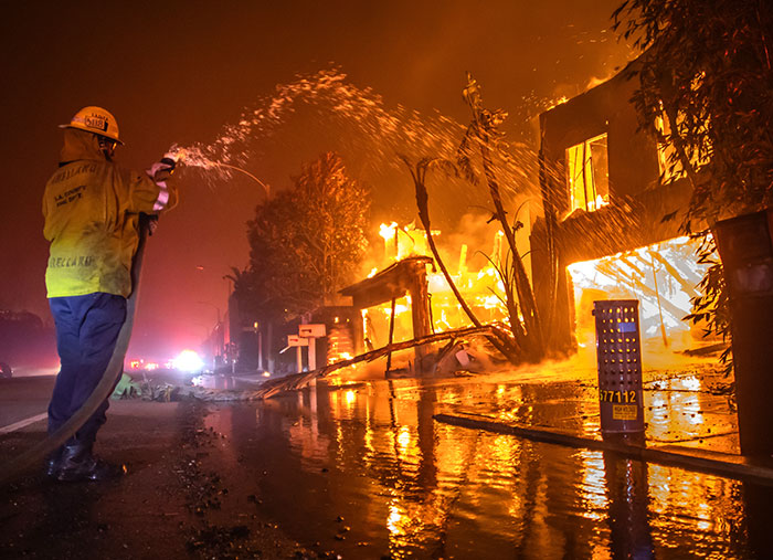 Firefighter battling a blaze during the LA fire, water spraying on burning structure at night.