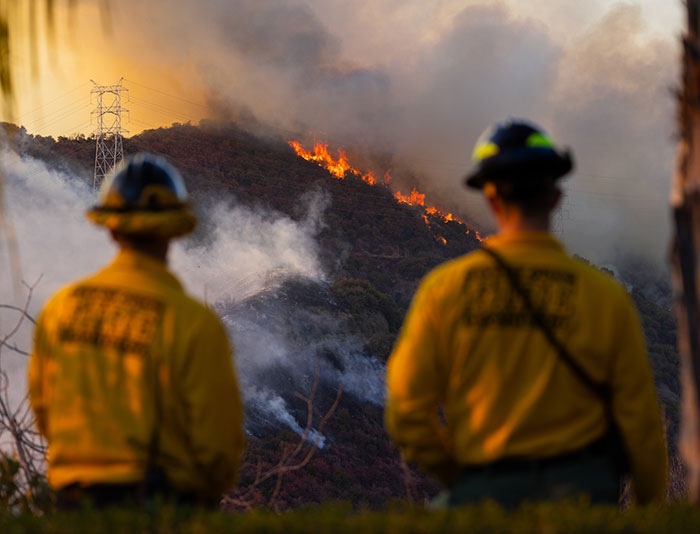 Private firefighters observe wildfire approaching a hillside, smoke and flames visible.