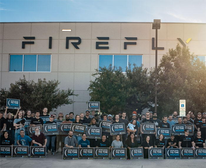 Group holding "To the Moon" signs in front of Firefly building, related to NASA's commercial robotic Moon launch.