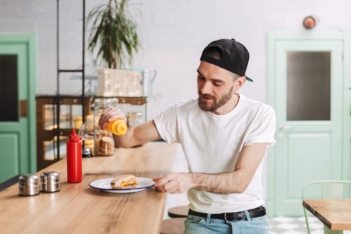 Man in a white shirt and black cap wasting food by adding excessive mustard to his meal at a dinner party.