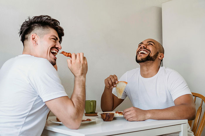 Two friends laughing while eating a home-cooked meal at a dining table.