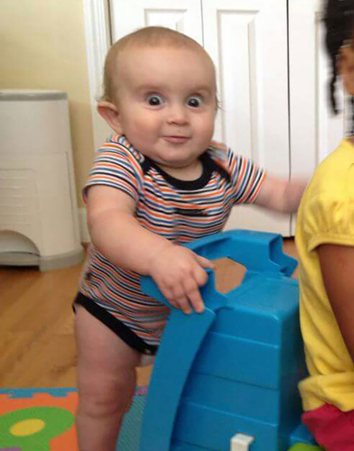 A baby makes a funny face while holding a blue toy, capturing a humorous moment for the camera roll.