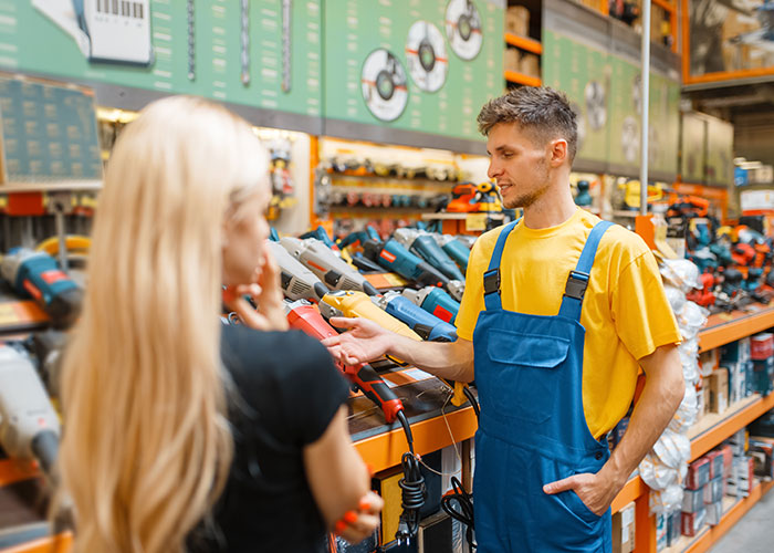 Man in blue overalls explaining power tools to a woman in a store, highlighting mansplaining dynamics in everyday situations.