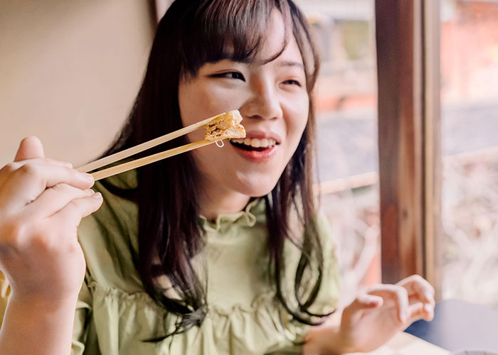 Woman smiling, holding food with chopsticks, embodying the concept of mansplaining in a relaxed setting.