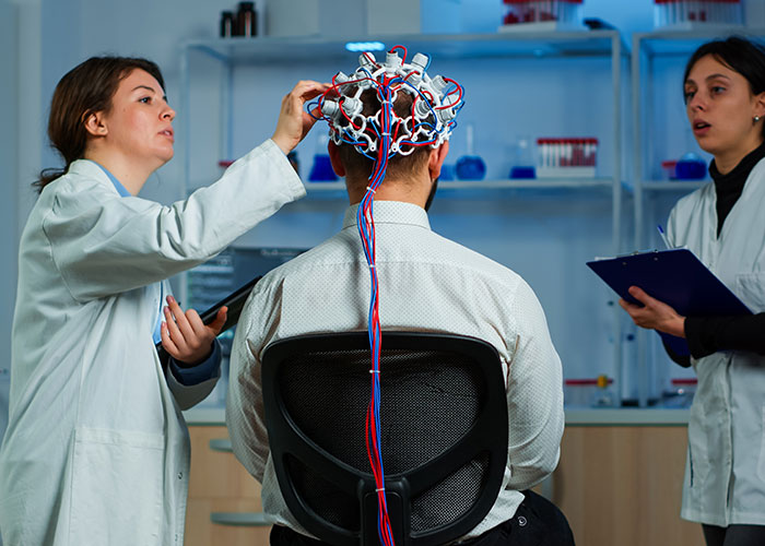 Two women in lab coats working with a man wearing an EEG cap in a laboratory setting.