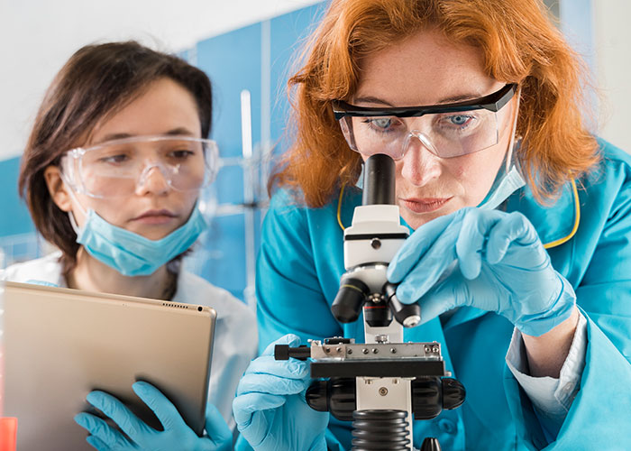 Two female scientists, one using a microscope, focusing intently, wearing protective goggles and lab coats.