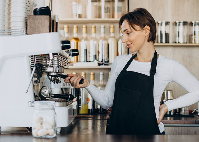 Barista in a cafe making coffee, smiling and wearing a black apron, surrounded by bottles and cups.